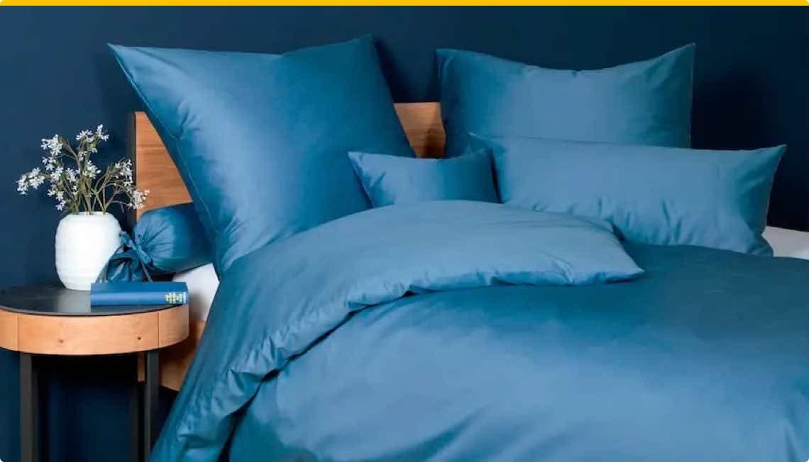 Bettwäsche-Set in Blau mit mehreren Kissen auf einem Bett. Blumen und ein Buch auf einem Nachttisch.