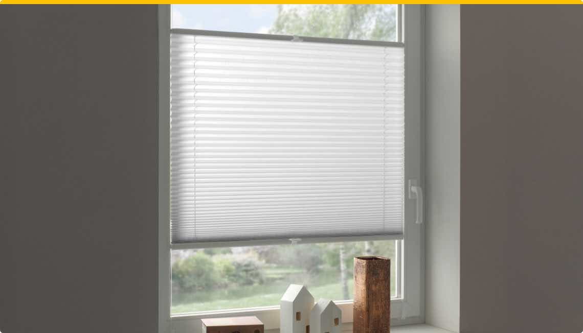 Weißes Plissee-Rollo an einem Fenster, darunter Dekoration mit kleinen Häuschen und einem Holzobjekt.