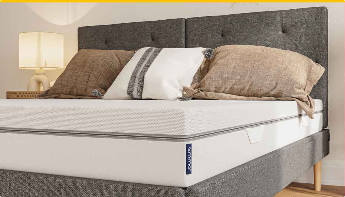 Gemütliches Bett mit grauem Kopfteil, weißer Matratze und braunen Kissen, neben einer Tischlampe.