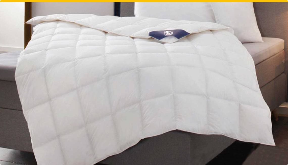 Weiße Steppdecke auf grauem Bett mit Kopfkissen im Hintergrund.