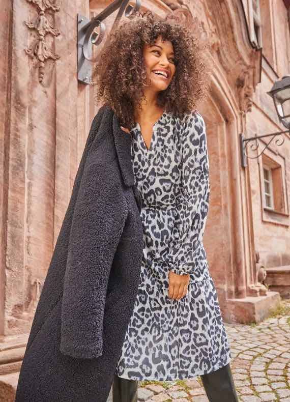 Frau in grauem Leopardenkleid hält einen schwarzen Mantel über der Schulter, steht vor einem historischen Gebäude.