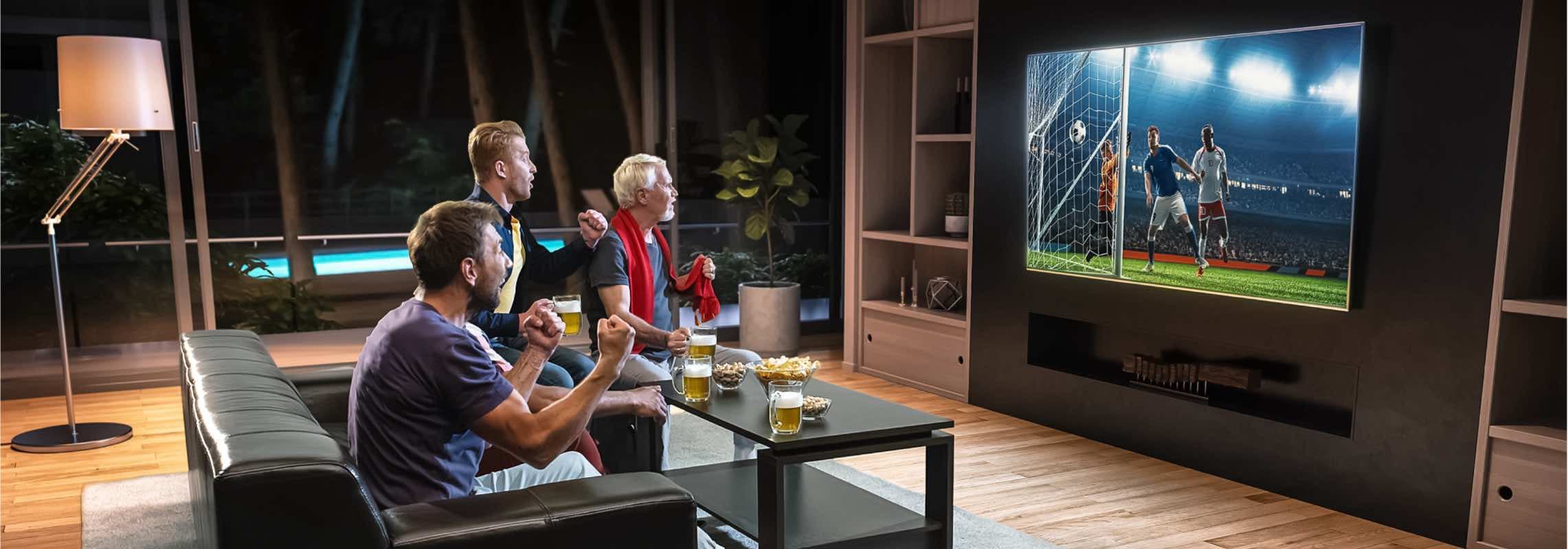 Drei Personen schauen Fußballspiel im Wohnzimmer, jubeln und trinken Bier. Fernseher zeigt Tor.