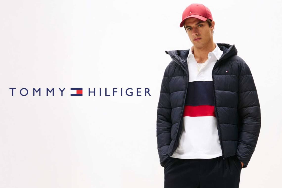 Tommy Hilfiger – Mann in gestreiftem Pullover und schwarzer Daunenjacke mit roter Kappe.