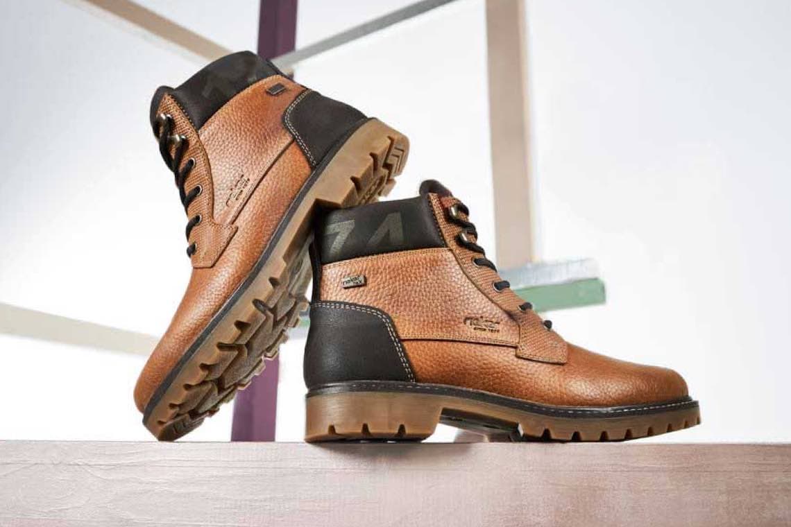 Zwei braune Stiefel mit schwarzem Absatz und robuster Sohle stehen auf einem Holztisch vor hellem Hintergrund.