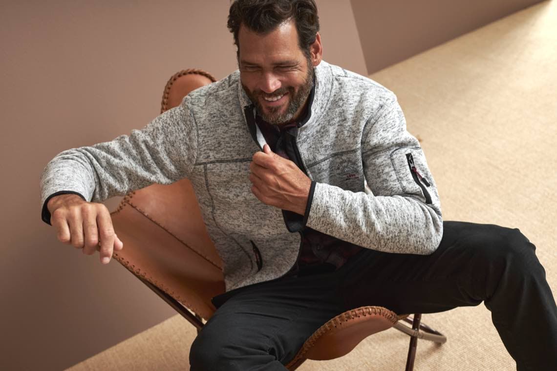 Mann sitzt auf einem Lederstuhl und trägt eine graue Jacke mit schwarzen Details.