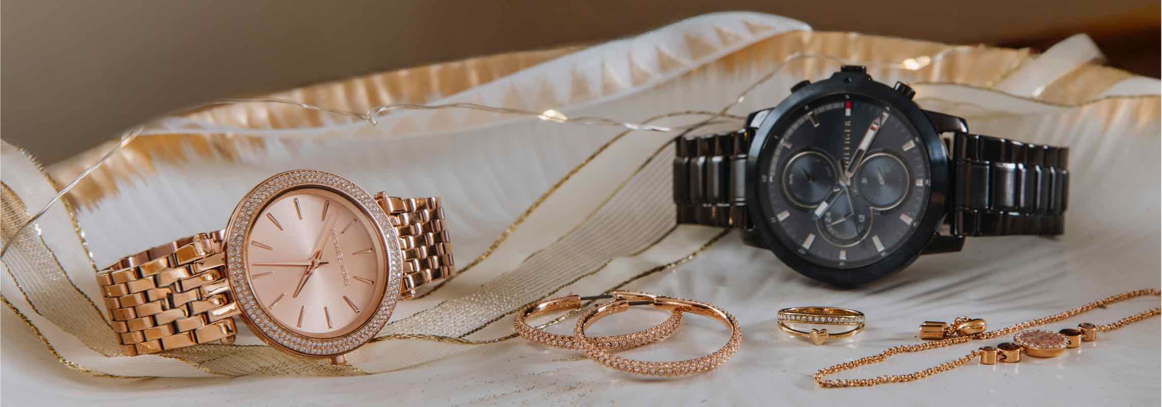 Zwei Armbanduhren, eine in Roségold und eine schwarze, umgeben von Schmuck auf einem dekorativen Hintergrund.