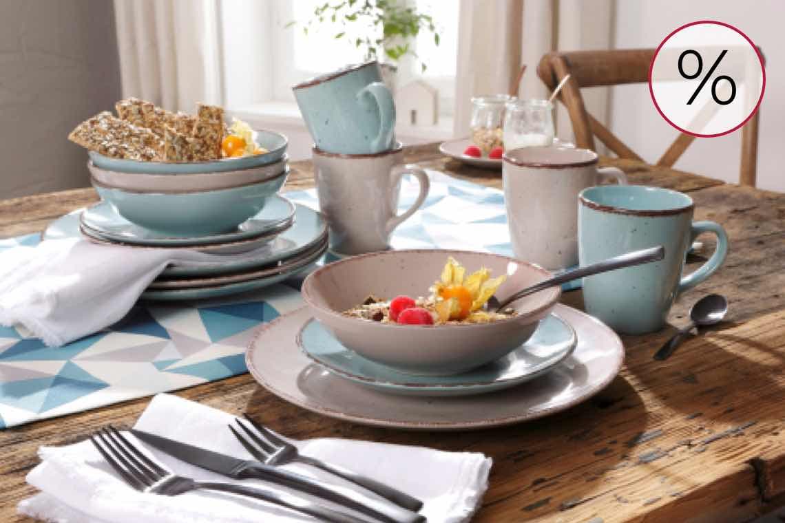 % – Frühstückstisch mit Geschirr und Müsli, Brotschale, Tassen und Besteck auf einem Holztisch