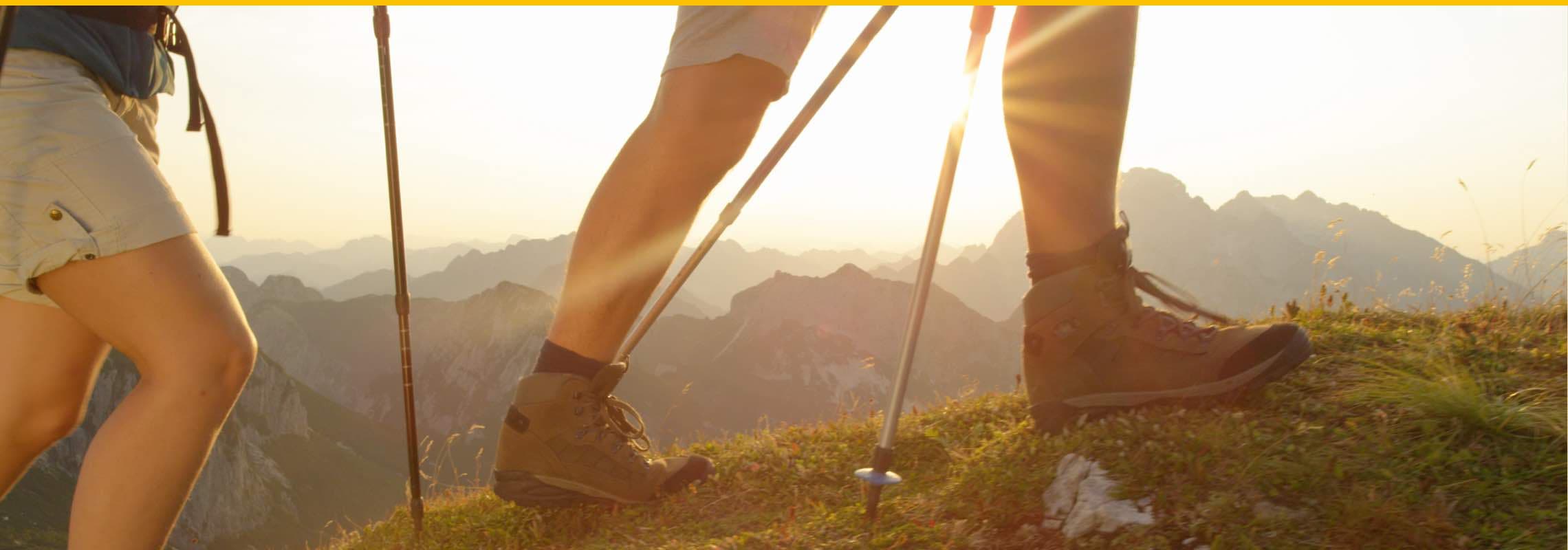 Zwei Personen wandern mit Wanderstöcken und Stiefeln bei Sonnenuntergang in den Bergen.
