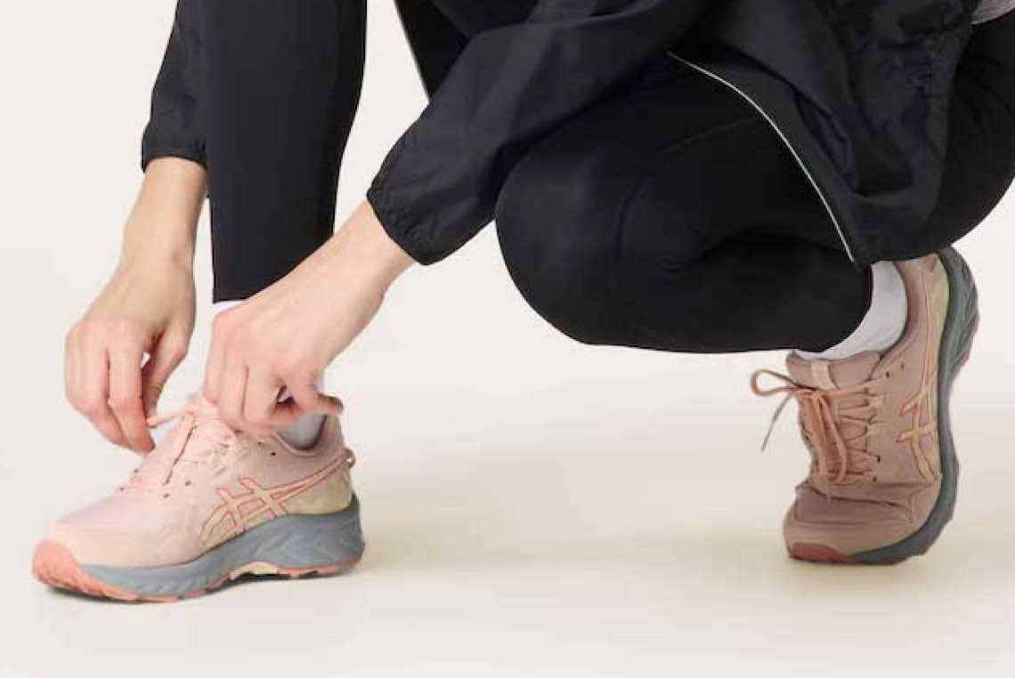 Person bindet Schnürsenkel an rosa Laufschuhen, kniet in schwarzer Sportkleidung auf dem Boden.