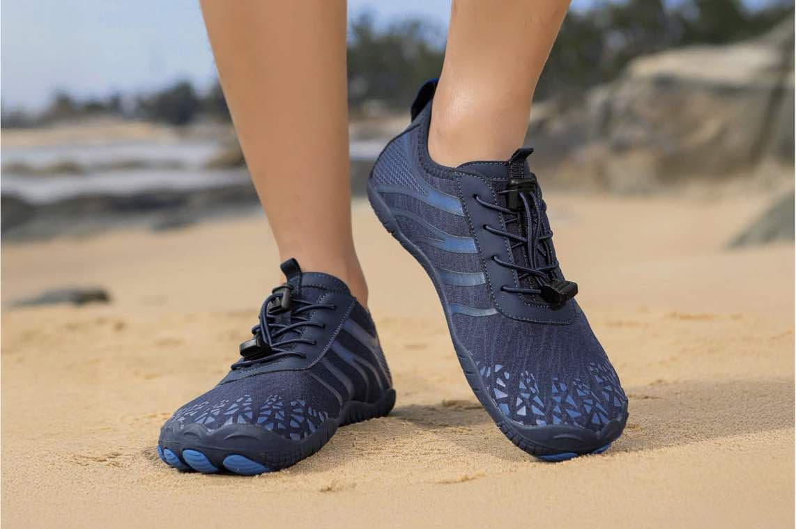 Dunkelblaue Sportschuhe mit Schnürsenkeln im Sand, Person trägt die Schuhe, Hintergrund mit Strand und Bäumen.