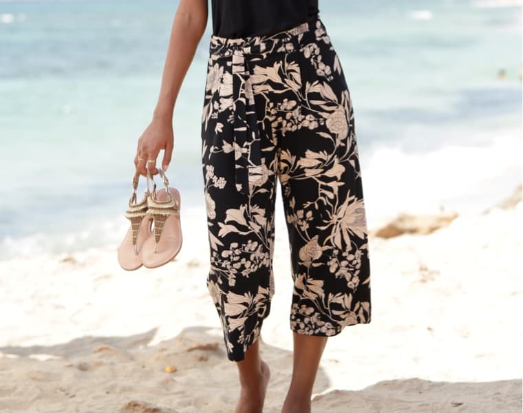 Person in geblümter Hose hält Sandalen in der Hand und steht am Strand.