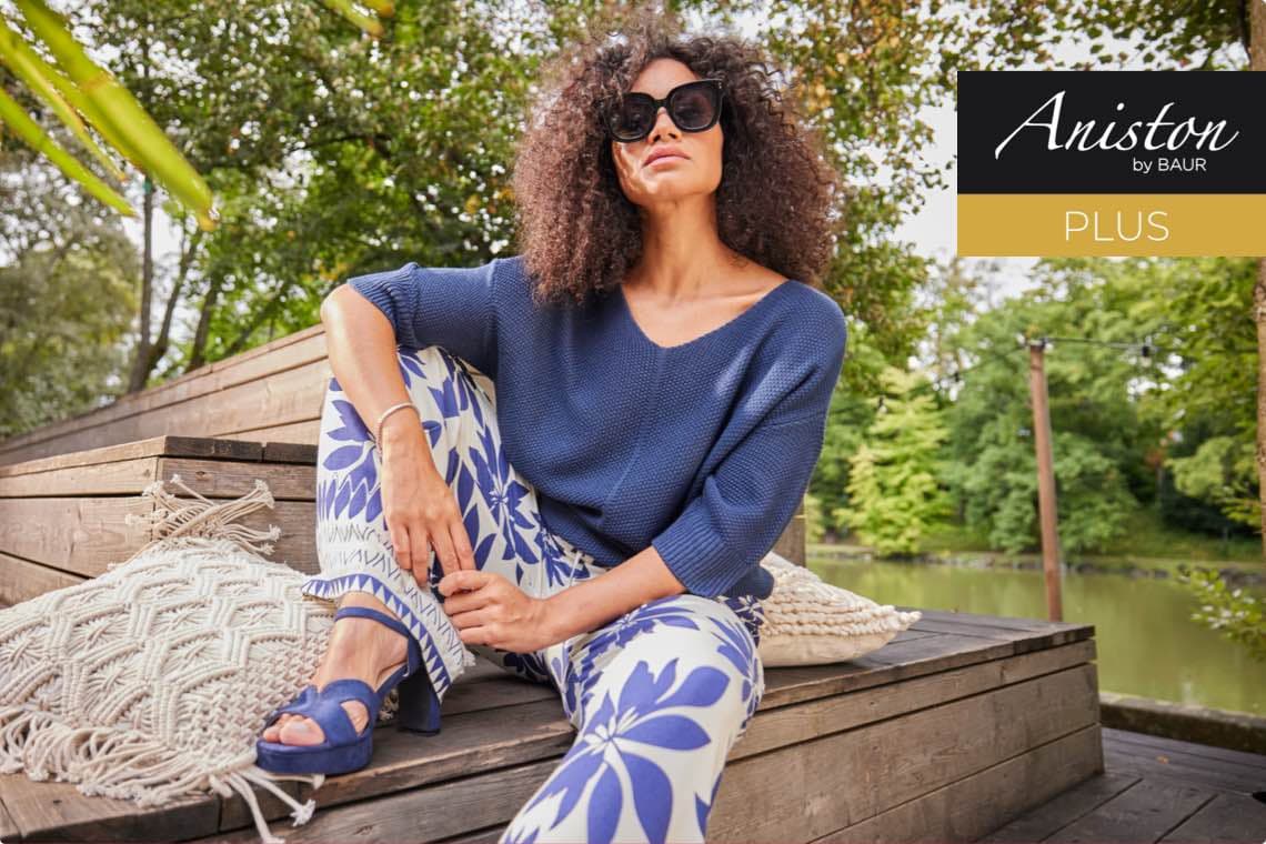 Aniston by Baur PLUS – Frau in blauem Pullover und gemusterter Hose sitzt auf Holzbank im Grünen