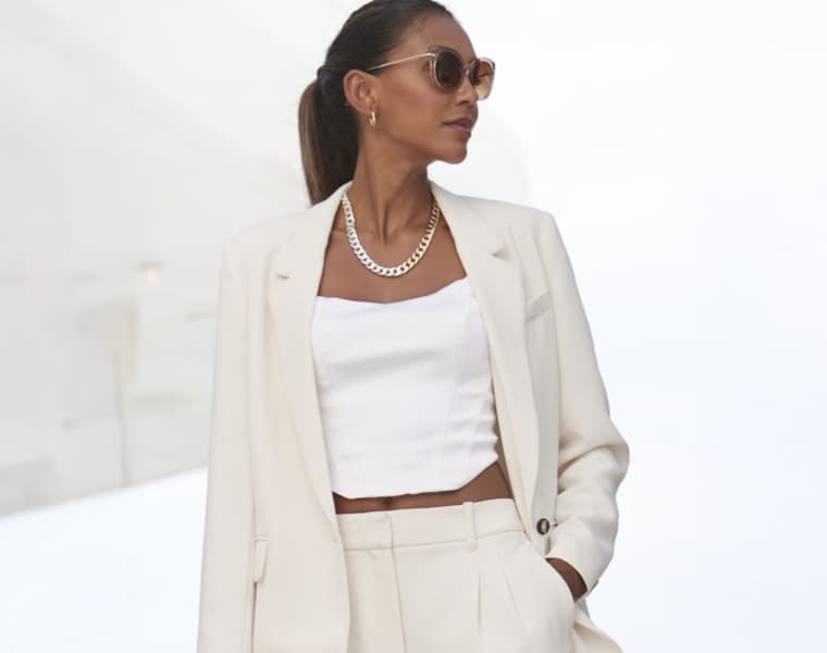 Frau in weißem Blazer, weißem Top und weißer Hose trägt eine Kette und hat die Hände in den Hosentaschen.