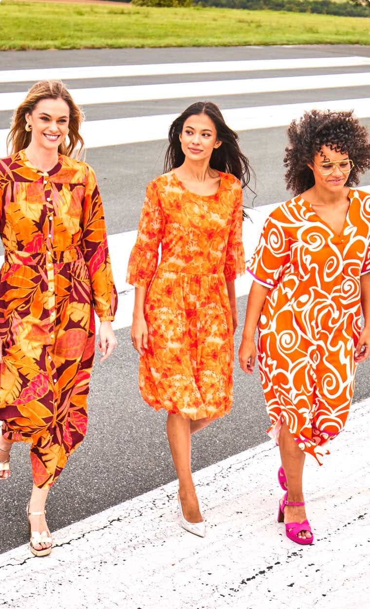 Drei Frauen in farbenfrohen Kleidern gehen nebeneinander über einen Zebrastreifen.