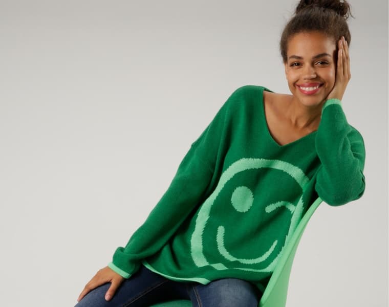 Person trägt grünen Pullover mit großem Smiley-Motiv und sitzt auf einem Stuhl.