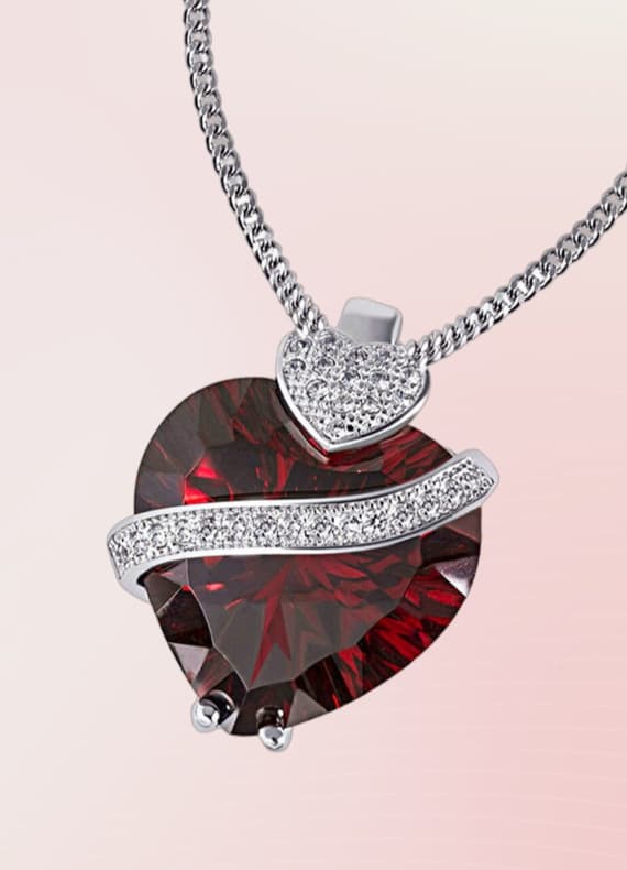 Herzförmiger Anhänger mit rotem Stein und glitzerndem Band auf silberner Kette vor rosa Hintergrund.