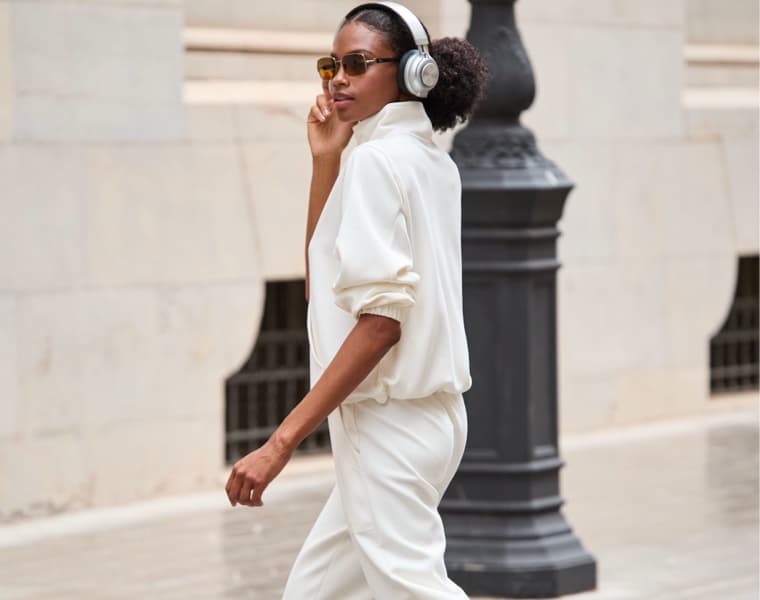 Person in weißem Outfit und Kopfhörern geht draußen entlang einer Steinmauer.