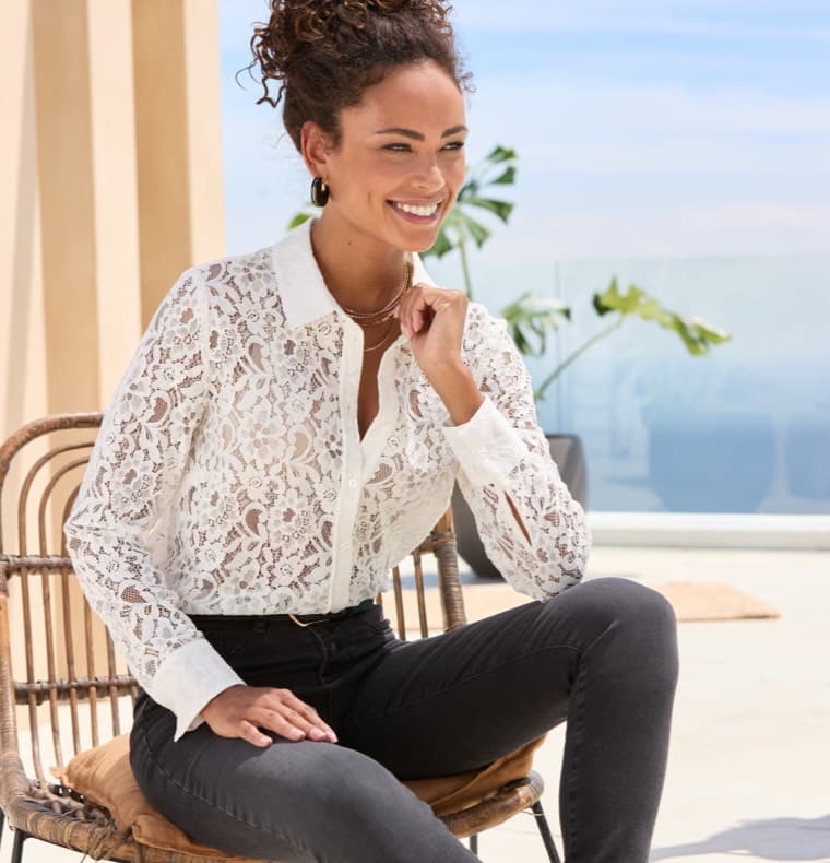 Frau in weißer Spitzenbluse und dunklen Jeans sitzt auf einem Stuhl, entspannter Outdoor-Hintergrund.