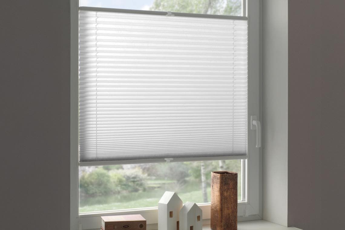 Geschlossene weiße Jalousie an einem Fenster, davor kleine Deko-Häuser und eine Holzsäule auf der Fensterbank.