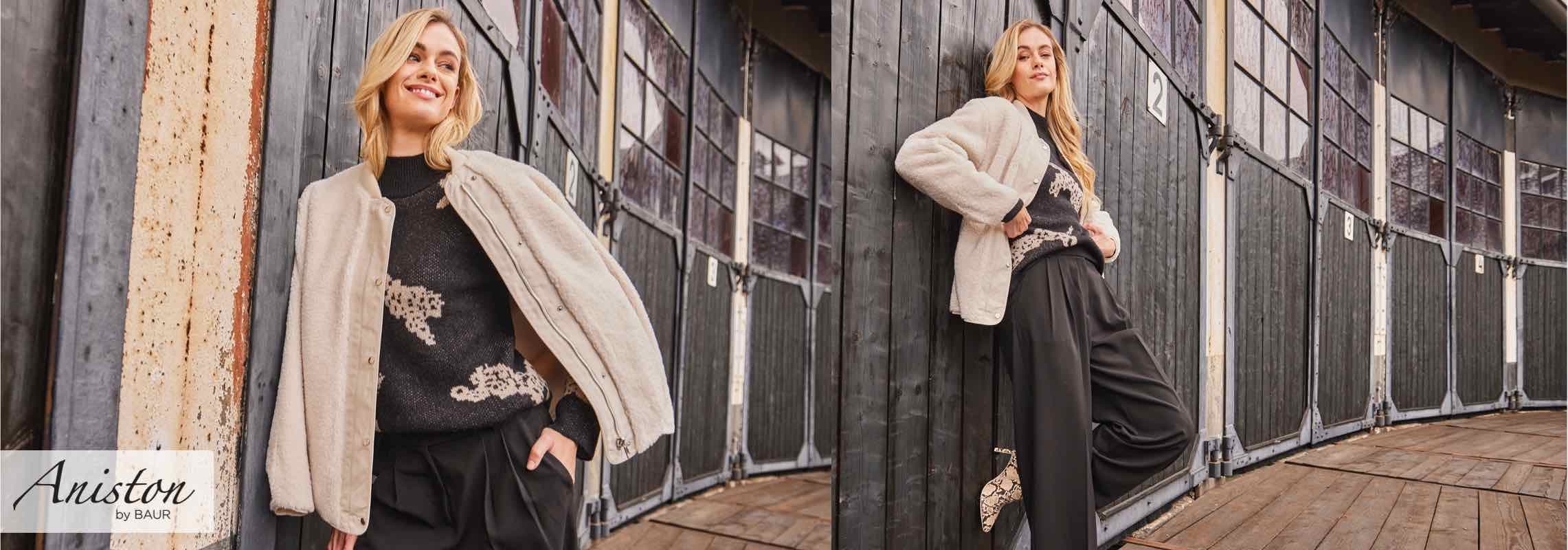 Aniston by BAUR – Frau lehnt an Holzwand, trägt beige Jacke und schwarzen Pullover mit Muster, dazu weite schwarze Hose und gemusterte Schuhe.