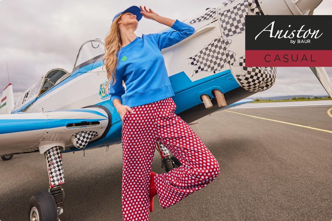 Aniston by BAUR Casual – Frau in blauem Pullover und roter Hose mit weißen Punkten vor Flugzeug.