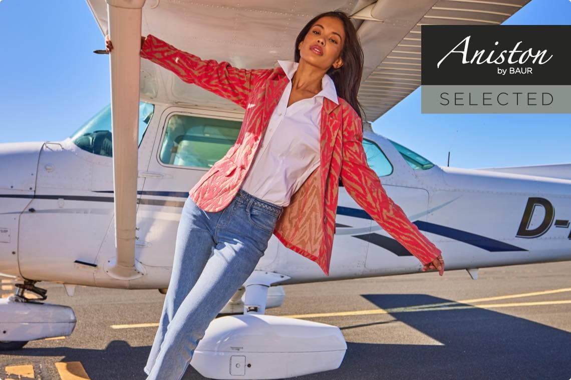 Aniston by BAUR SELECTED – Frau in rotem Blazer lehnt sich an ein Flugzeug auf dem Rollfeld.