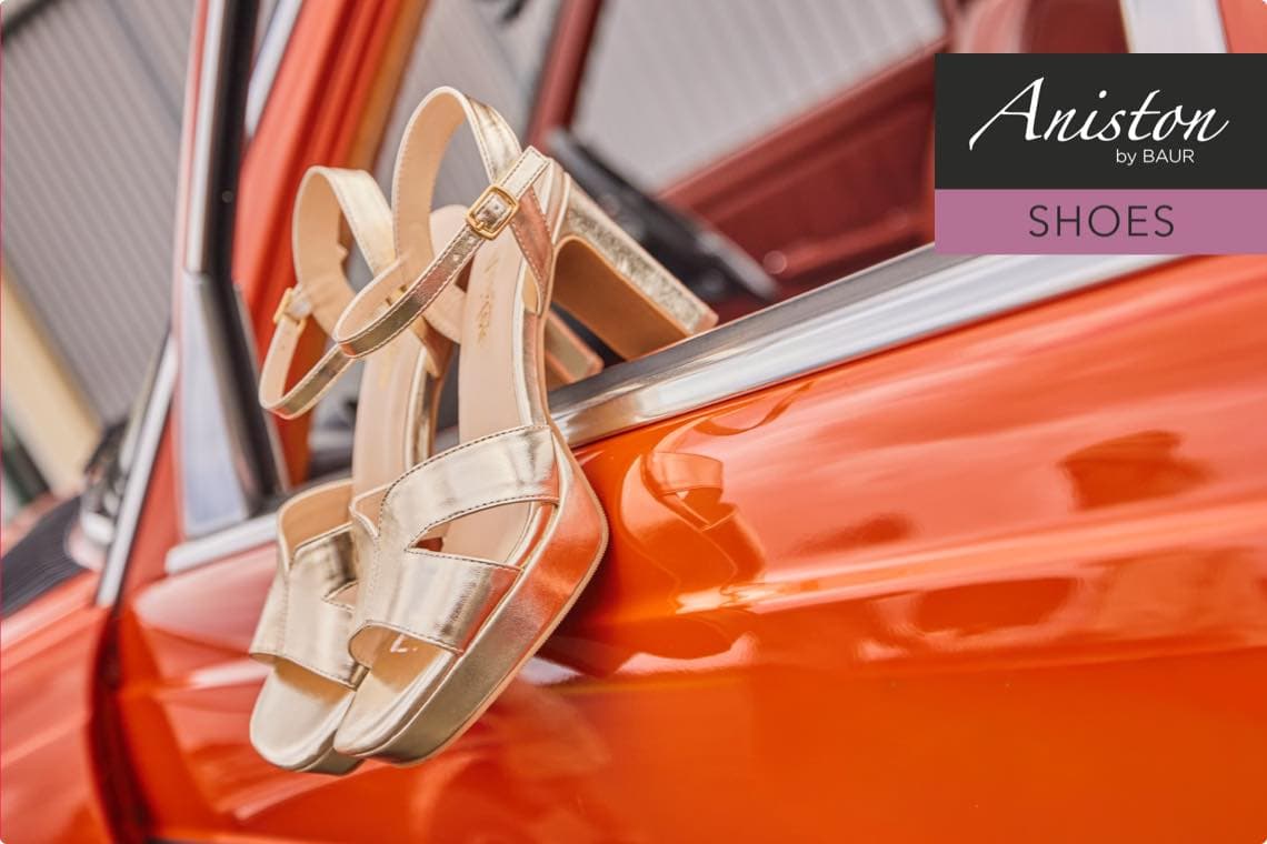 Aniston by Baur Shoes – Goldene Sandaletten hängen an einem orangefarbenen Autofenster.