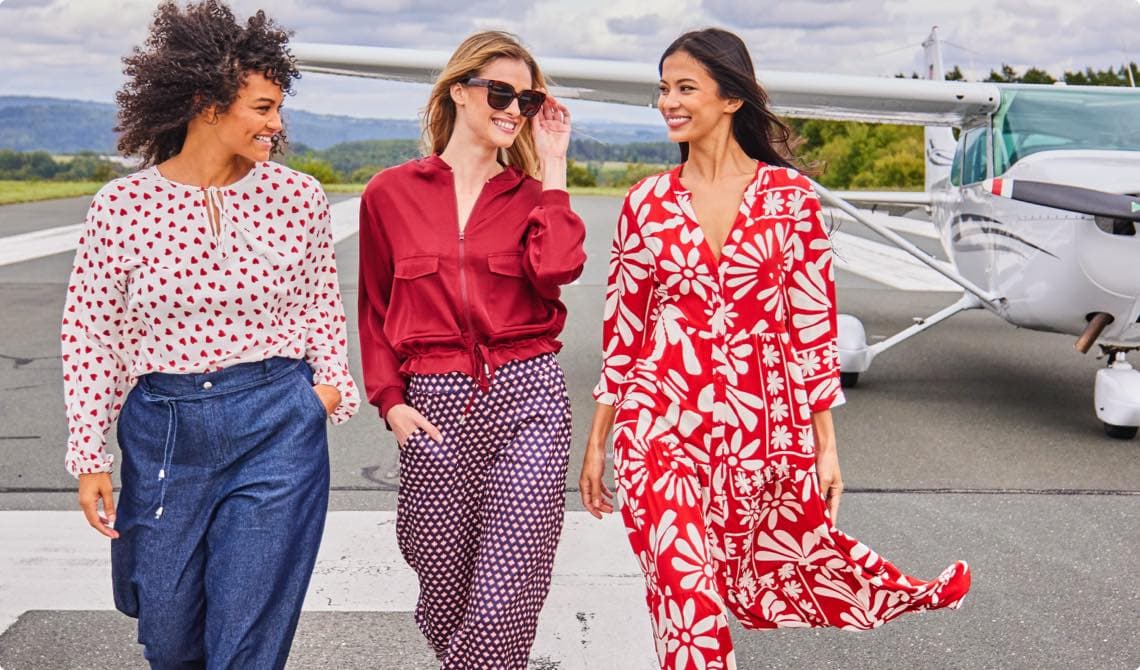 Drei Frauen in modischer Kleidung gehen auf einer Landebahn, ein Flugzeug im Hintergrund.