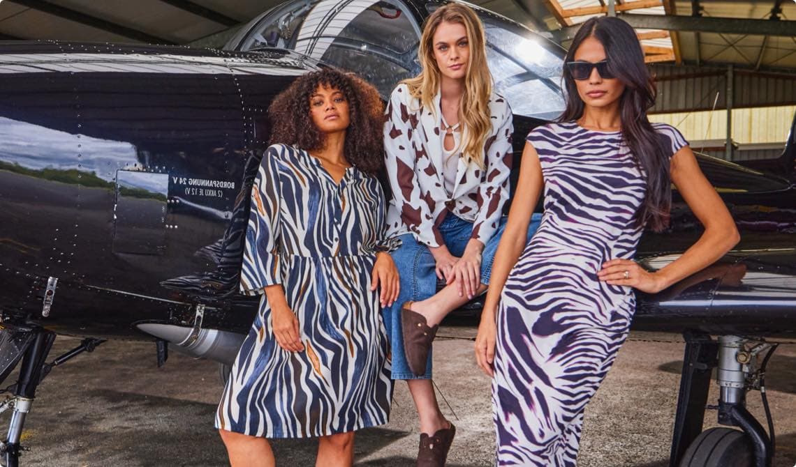 Drei Frauen in gemusterten Outfits posieren vor einem schwarzen Flugzeug in einem Hangar.