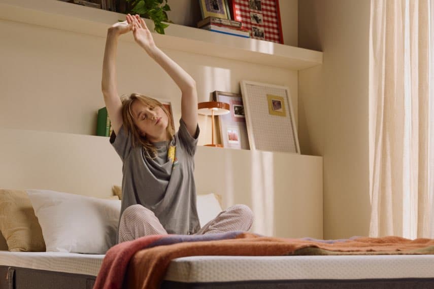 Frau sitzt auf einem Bett und streckt sich, Schlafzimmer mit Regalen und Dekoration im Hintergrund.