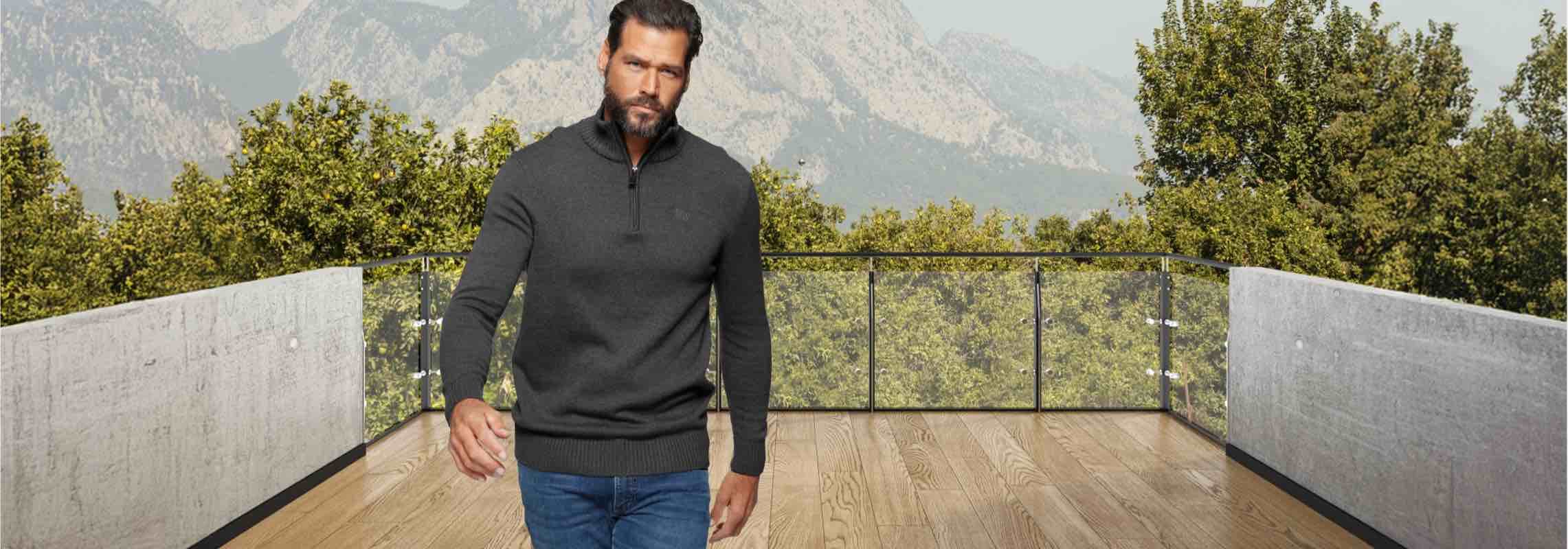 Mann in grauem Pullover und blauer Jeans geht auf einer Terrasse, Hintergrund mit Bergen und Bäumen.