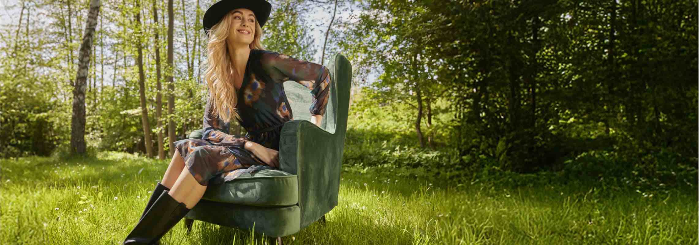 Frau mit schwarzem Hut und transparentem Kleid sitzt auf einem grünen Sessel im Wald.