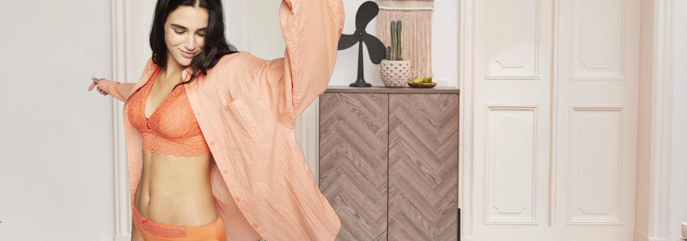 Frau in korallfarbener Unterwäsche und passendem Hemd posiert vor einem Schrank mit dekorativen Gegenständen.