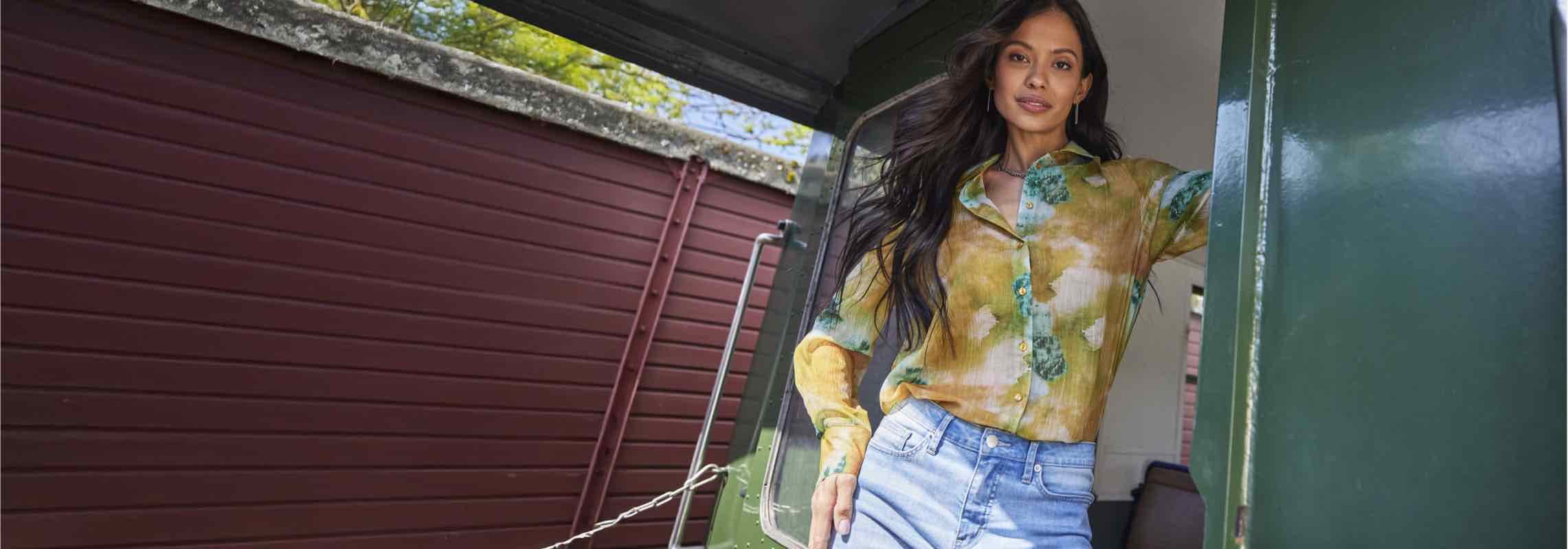 Frau in gelbem, gemustertem Hemd und blauer Jeans steht in Türöffnung.