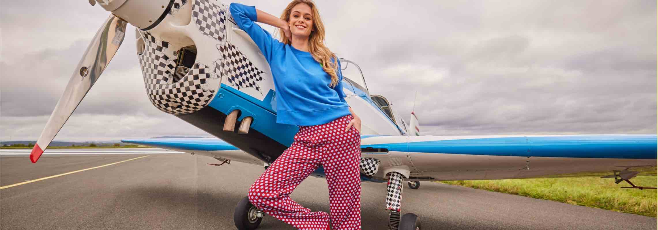 Frau in blauem Pullover und roter Hose lehnt sich gegen ein blau-weißes Flugzeug auf einer Rollbahn.