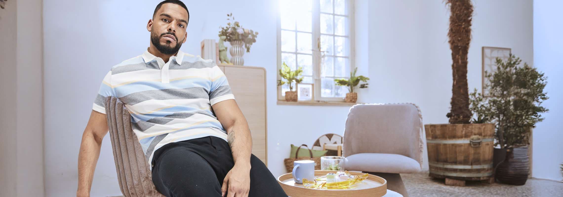 Mann in gestreiftem Poloshirt sitzt entspannt auf einem Stuhl in einem hellen Zimmer mit Pflanzen.
