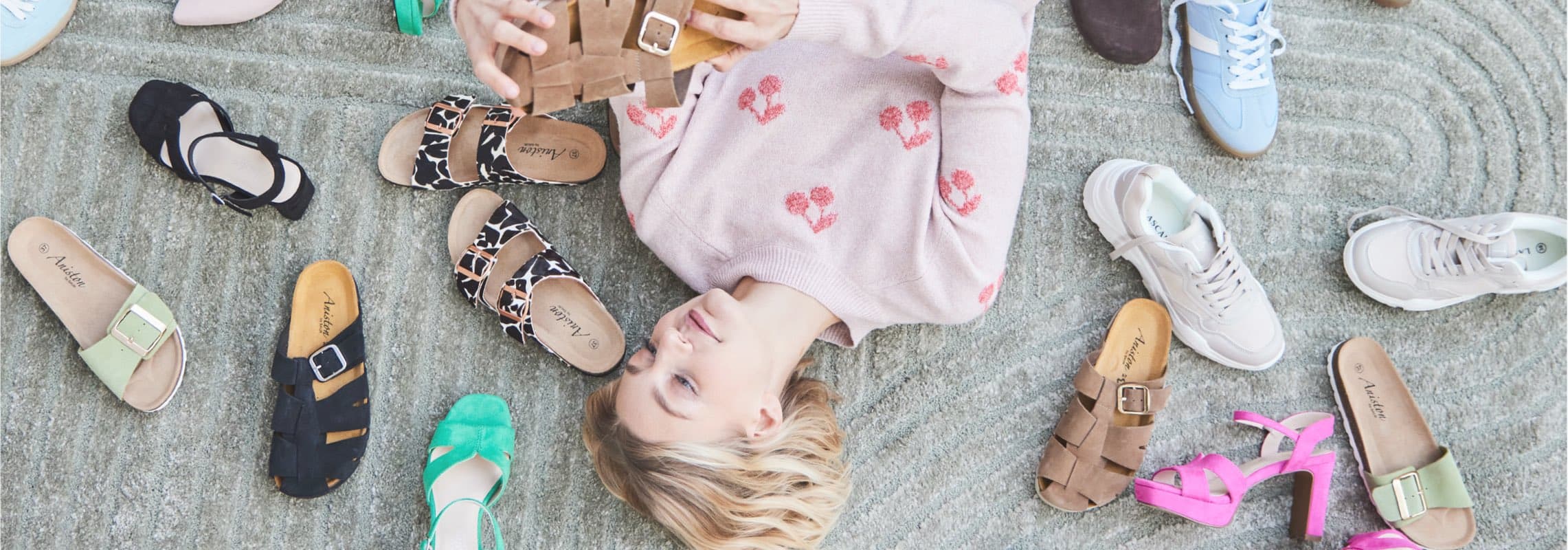 Person im rosa Pullover liegt auf Teppich, umgeben von verschiedenen Schuhen in bunten Farben.