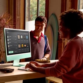 Zwei Personen an einem Schreibtisch, eine arbeitet an einem PC, die andere steht daneben und schaut zu.