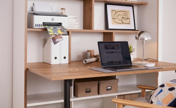 Modern Arbeitsbereich mit Laptop, Drucker, Regalen und Lampe auf Holzschreibtisch, gemütlicher Stuhl daneben.