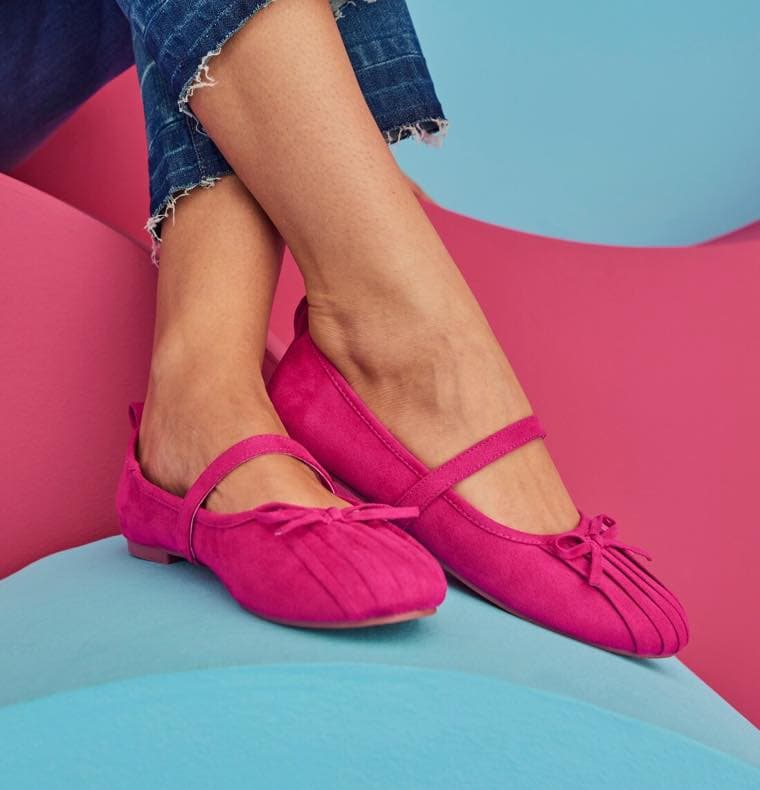 Person trägt pinke Ballerinas mit Schleifen, gekreuzte Beine, Jeans mit ausgefranstem Saum, bunte Hintergrundfarben