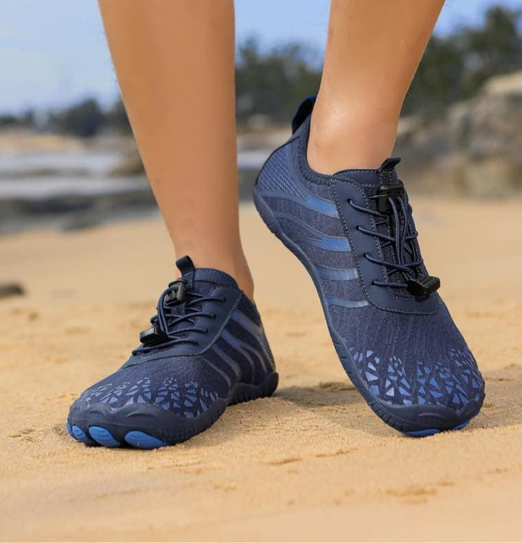Dunkelblaue sportliche Barfußschuhe im Sand getragen, Nahaufnahme der Füße mit Strand im Hintergrund.