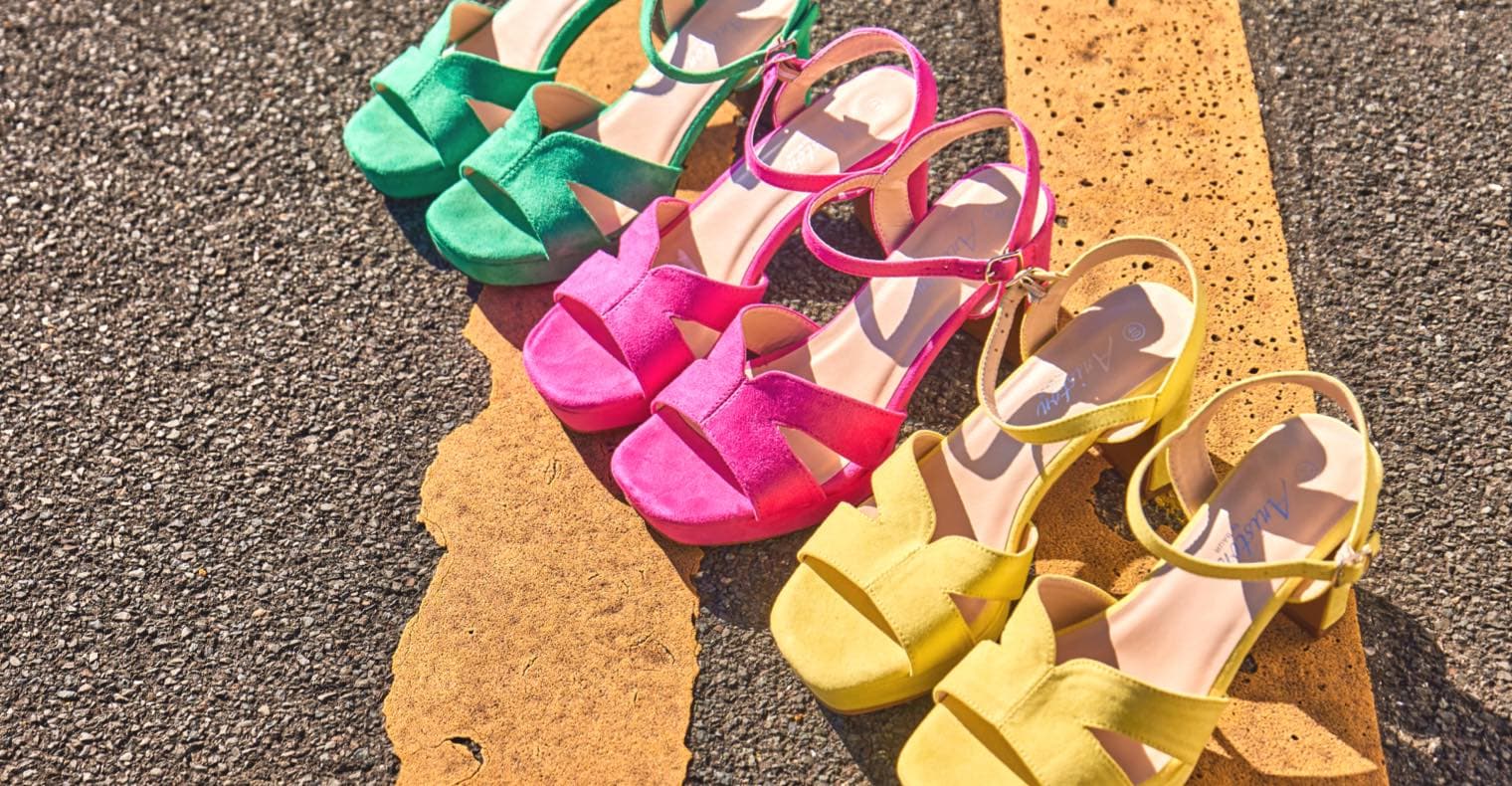 Vier Paar Sandalen in Grün, Pink, Gelb und Beige nebeneinander auf Asphalt und gelber Markierung.