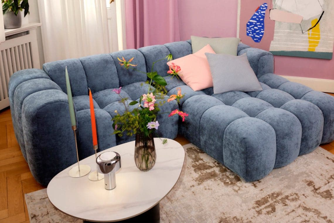Blaue modulare Couch mit bunten Kissen in gemütlichem Wohnzimmer, Blumen und Kerzen auf modernem Couchtisch.