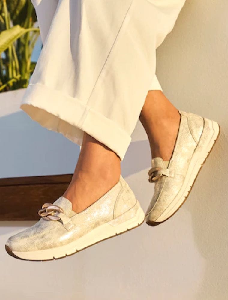 Füße in hellen Slippern mit dekorativer Kette, getragen zu heller weiter Hose vor sonniger Wand