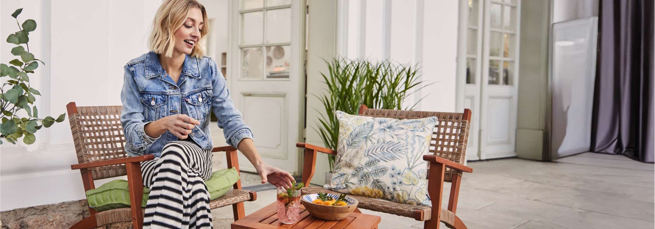 Frau in Jeansjacke und gestreifter Hose sitzt draußen, greift nach einem Getränk auf einem Tisch. Ein Kissen auf einem Stuhl.