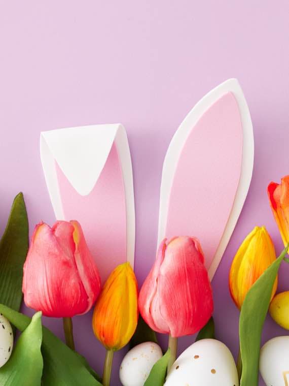 Osterdekoration mit Tulpen, Ostereiern und rosa Hasenohren vor einem lila Hintergrund