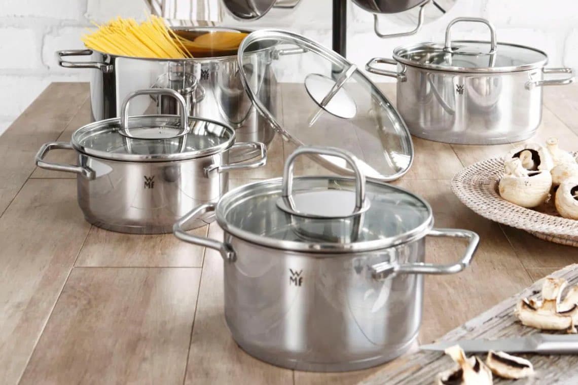 Mehrere Edelstahlkochtöpfe auf Holztisch, daneben Pilze und Nudeln in einem Topf.