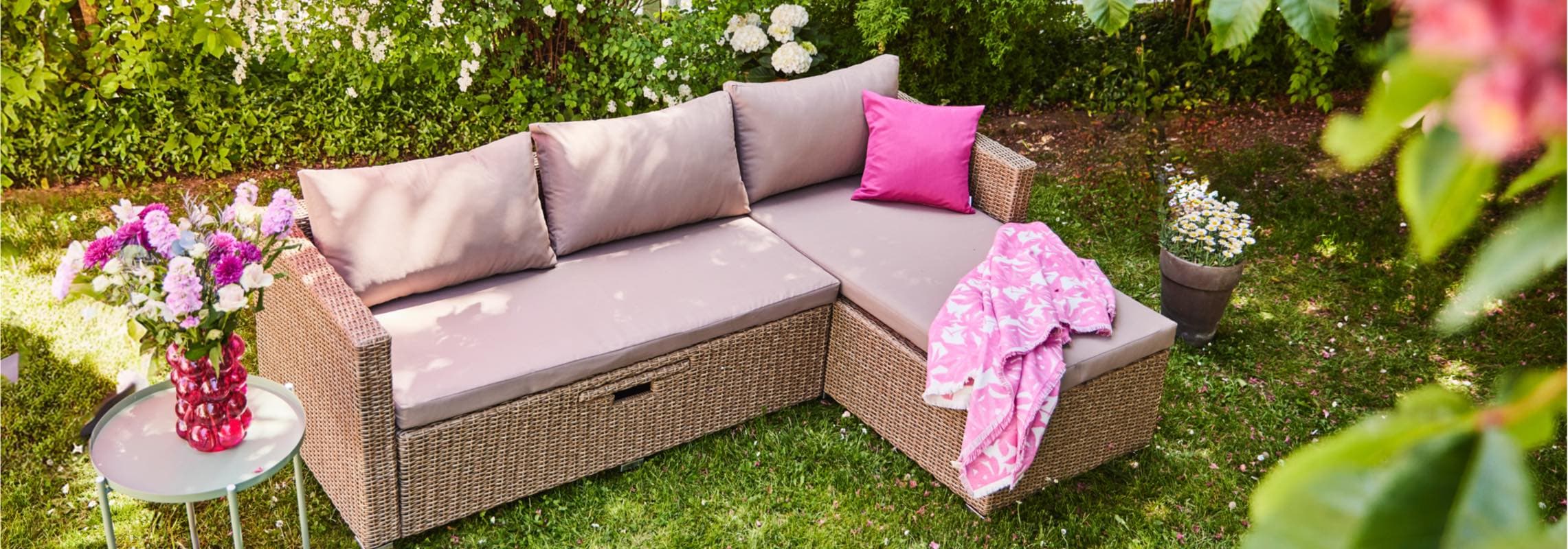 Gartensofa mit beigen Kissen und pinkem Kissen, daneben Tisch mit Blumenvase, umgeben von grüner Hecke und Blumen.