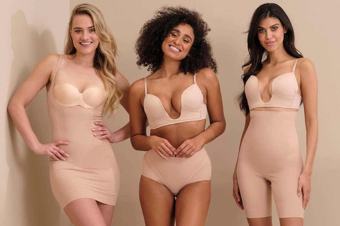 Drei Frauen in hautfarbenen Shapewear-Outfits posieren vor beigem Hintergrund.