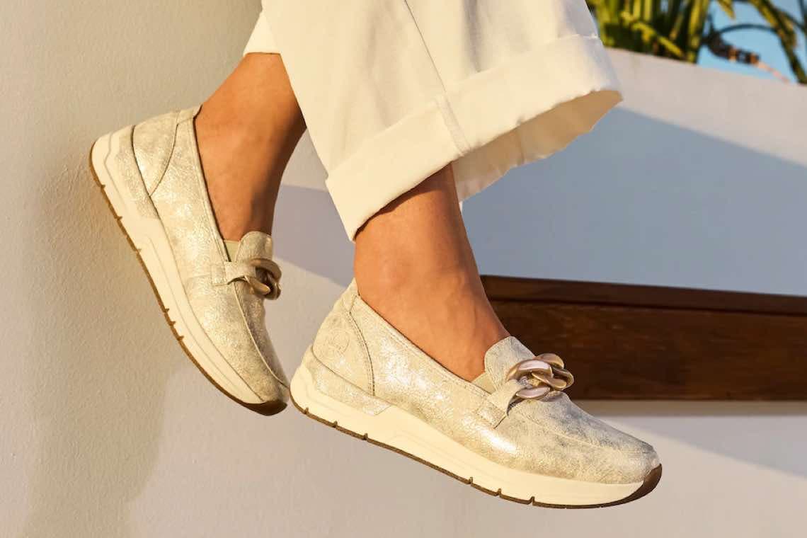 Person trägt goldene Loafers und weiße Hose vor hellem Hintergrund.